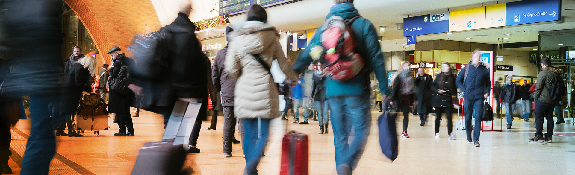 Reisende mit Gepäck in einer belebten Bahnhofshalle.
