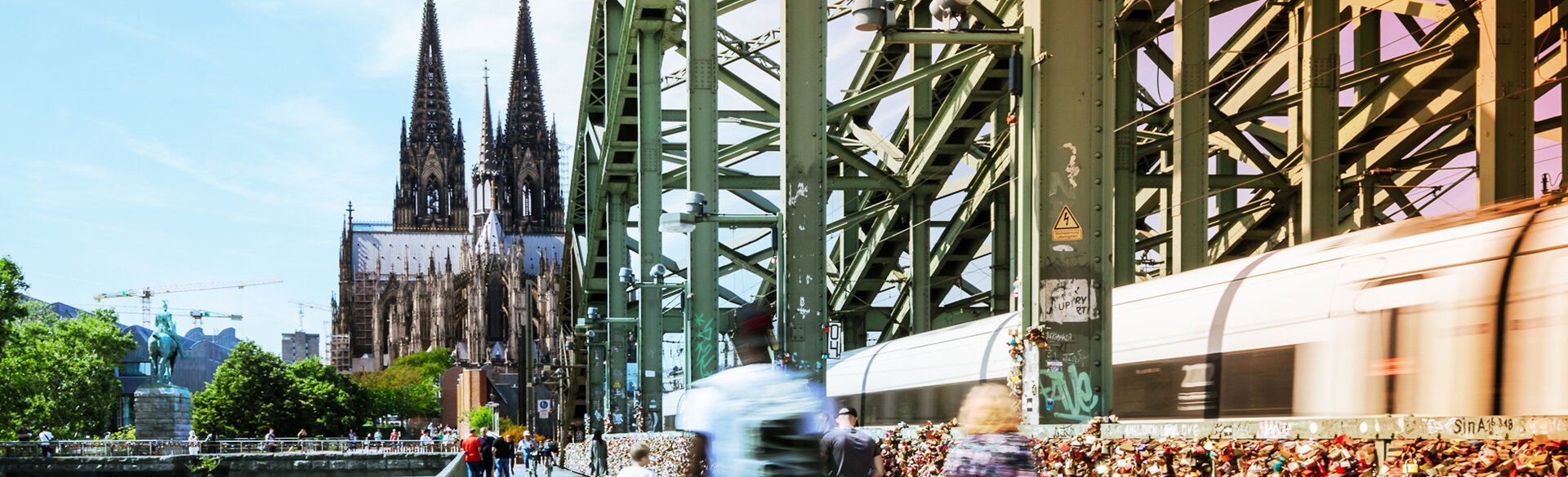 Stadtansicht von Köln mit dem Kölner Dom und einer Stadtbahn auf einer Brücke.