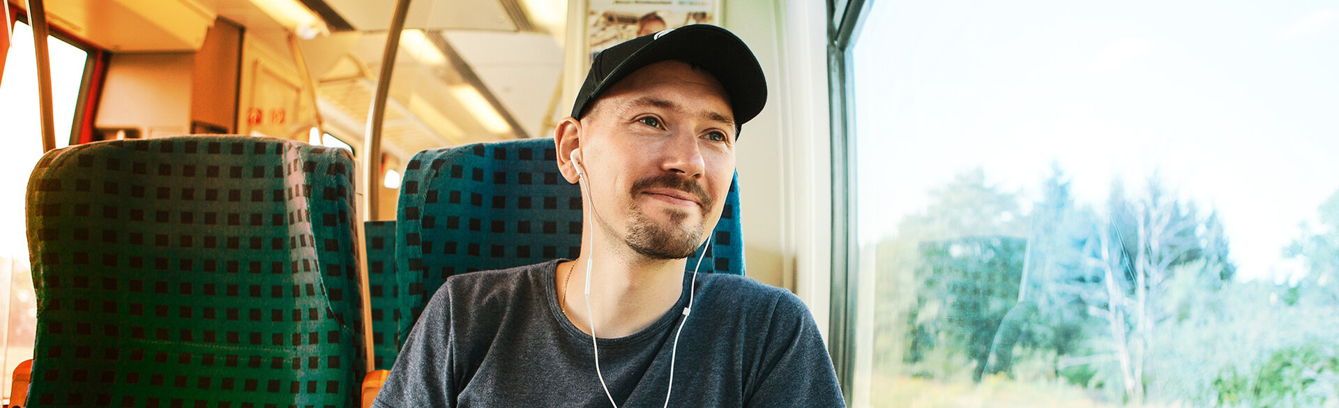 Ein Mann mit schwarzer Kappe und Kopfhörern sitzt am Fenster eines Zuges und blickt nach draußen.