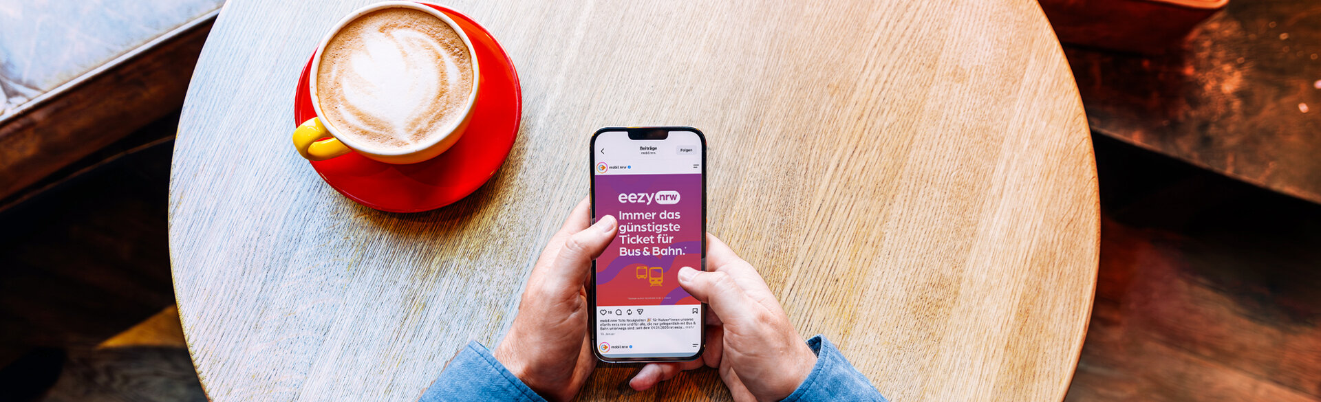 Blick von oben auf eine Person, die an einem Holztisch ein Smartphone in den Händen hält. Auf dem Display ist ein Beitrag zu eezy.nrw mit dem Schriftzug „Immer das günstigste Ticket für Bus & Bahn“ zu sehen. Daneben steht eine Tasse Kaffee auf dem Tisch.