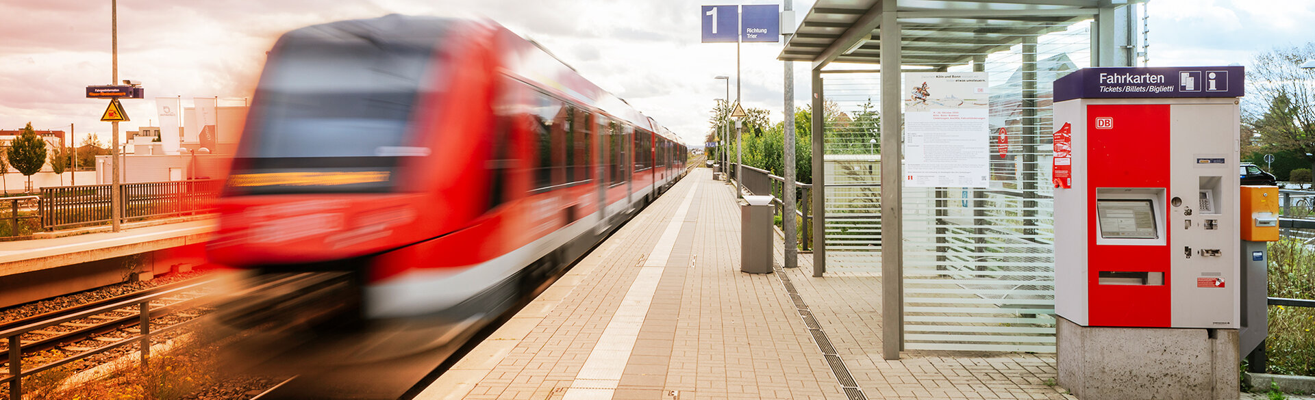 Ein roter Regionalzug fährt an einem Bahnsteig ein. Rechts befindet sich ein Fahrkartenautomat unter einem Wartehäuschen.