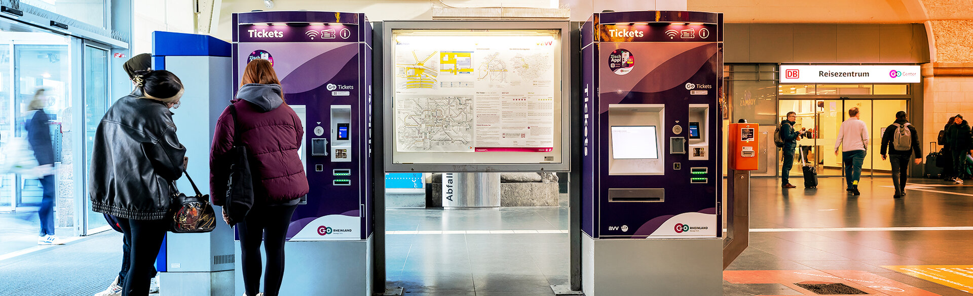 Zwei Fahrgäste stehen vor Ticketautomaten in einem Bahnhof. Im Hintergrund befinden sich weitere Bahnsteige und Reisende.