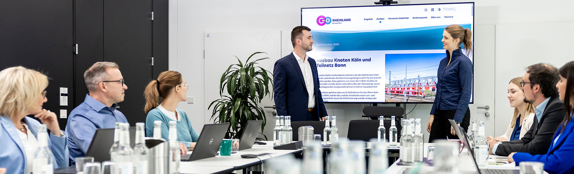 Besprechung in einem Konferenzraum mit mehreren Personen an einem Tisch und einem Vortragenden vor einem Bildschirm.