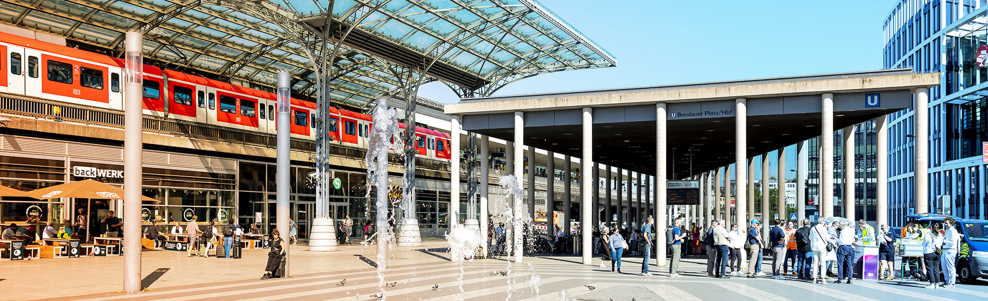 Blick auf den Breslauer Platz am Kölner Hauptbahnhof mit einer roten S-Bahn auf der Hochtrasse und dem Eingang zur U-Bahn-Station Breslauer Platz/Hbf. Der zentrale Umsteigepunkt verknüpft verschiedene Angebote des öffentlichen Nahverkehrs.