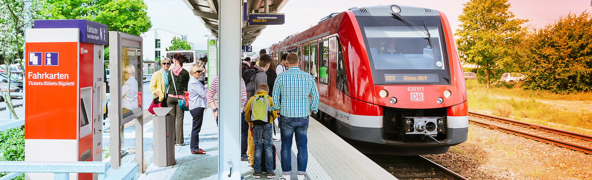 Fahrgäste an einem Bahnsteig neben einem roten Regionalzug und einem Fahrkartenautomaten.