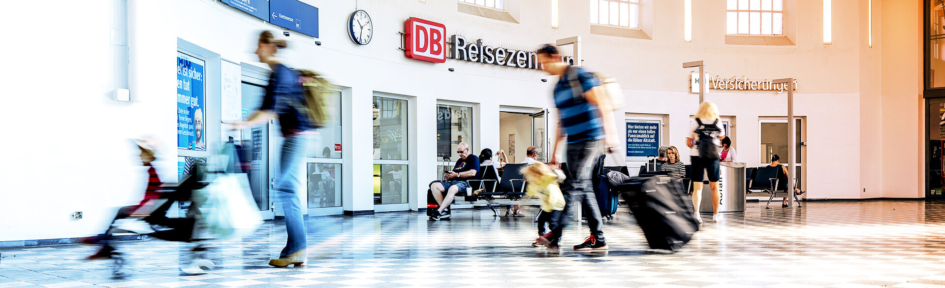 Bahnhofshalle mit Reisenden vor einem DB-Reisezentrum. Mehrere Personen bewegen sich mit Gepäck durch den Wartebereich.