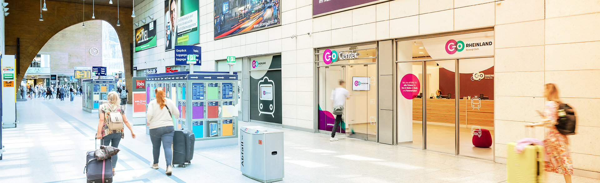 Reisende mit Gepäck in einer Bahnhofshalle. Rechts befindet sich ein Kundencenter von go.Rheinland.