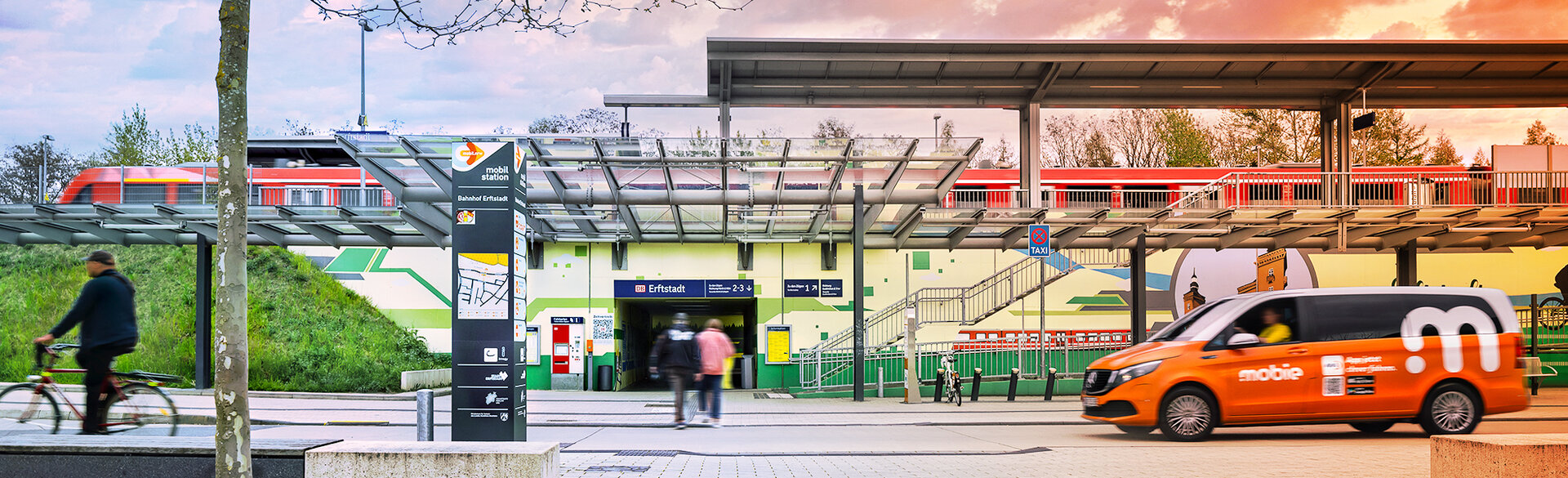 Ein Bahnhof mit Bahnsteigen und Fahrgastinformationen. Im Vordergrund fährt ein orangefarbener On-Demand-Kleinbus vorbei, links ist eine Person auf einem Fahrrad zu sehen.