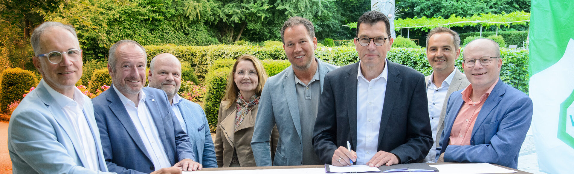 Gruppe von acht Personen steht im Freien um einen Tisch, auf dem Dokumente unterzeichnet werden.