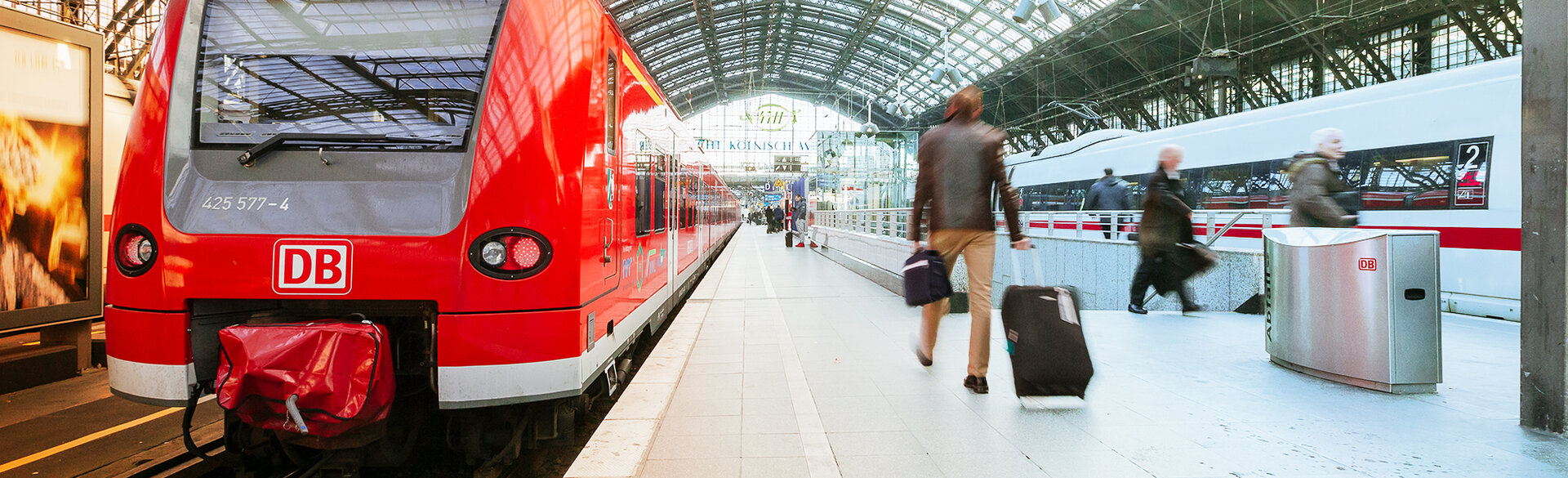 Roter Regionalzug und weißer ICE an den Bahnsteigen im Kölner Hauptbahnhof. Reisende mit Gepäck bewegen sich zwischen den Zügen. Das Motiv zeigt den Kölner Hauptbahnhof als zentralen Verknüpfungspunkt von Regional- und Fernverkehr.